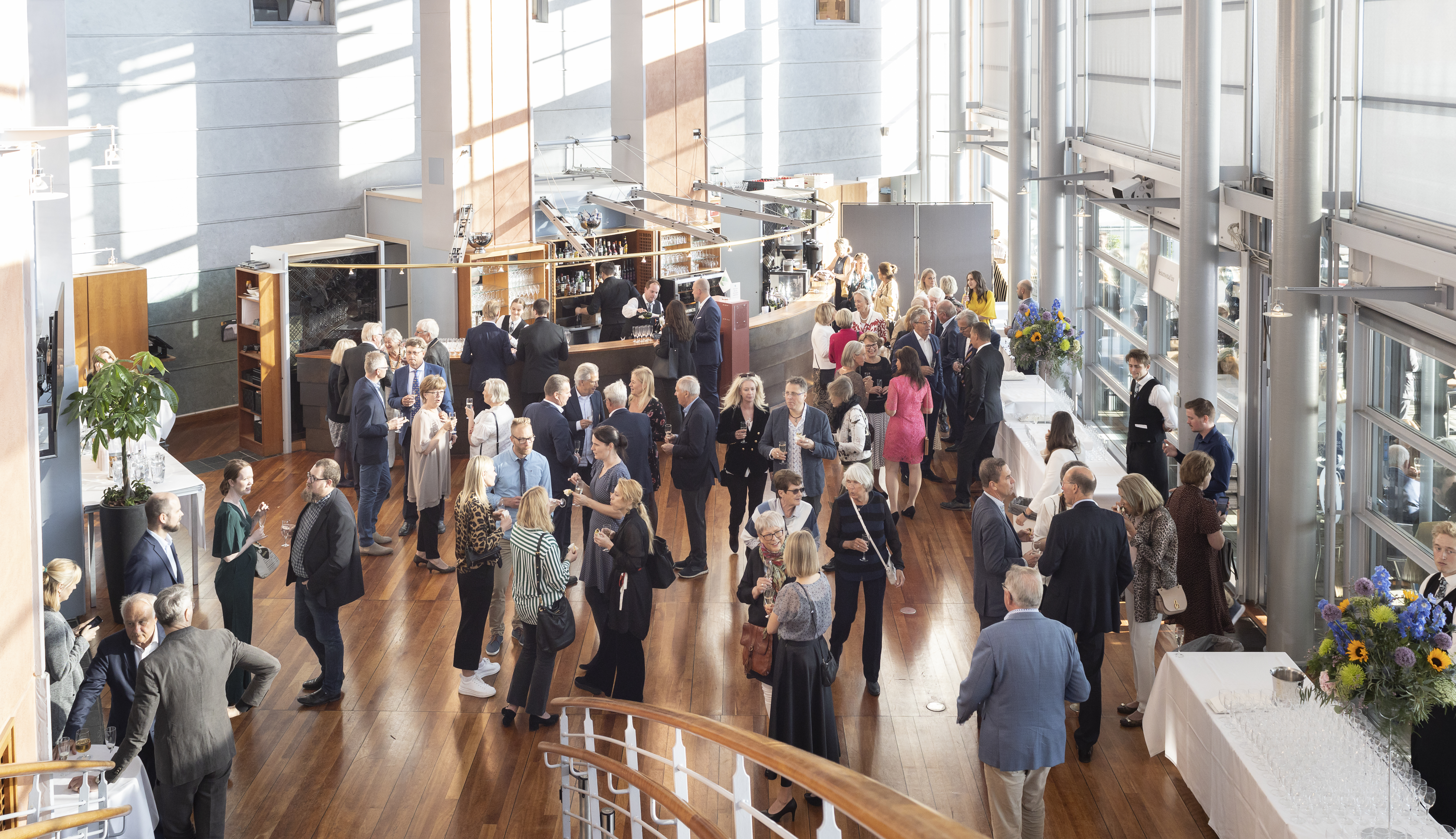 Många människor minglar i en väl upplyst teaterfoajé, nära en bar – sett uppifrån.