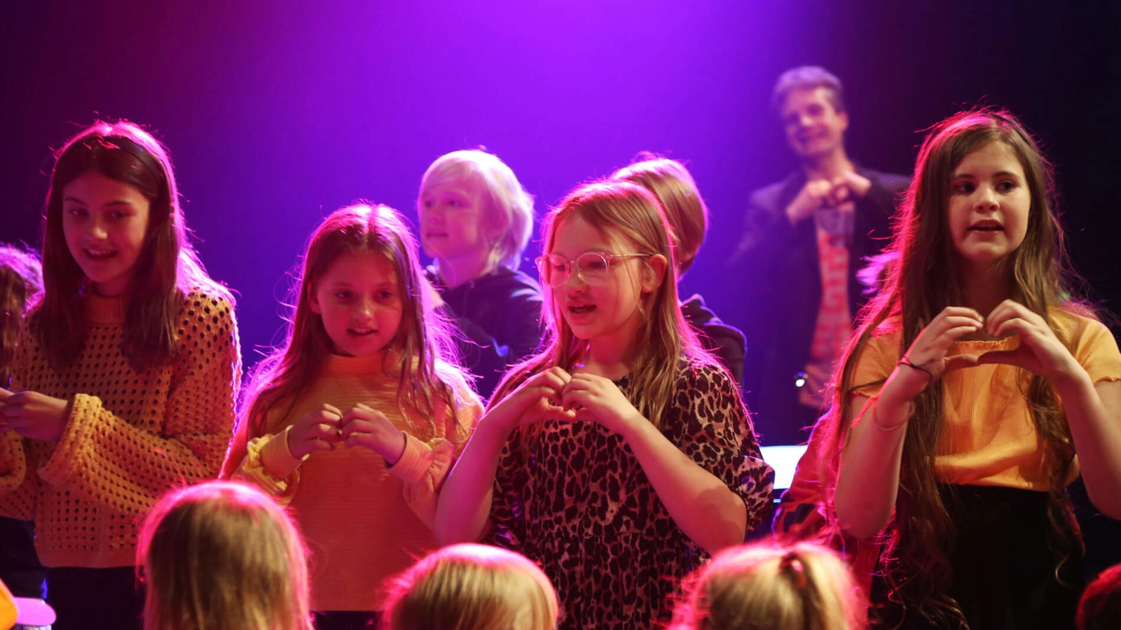 En barnkör sjunger, upplysta av lila och rosa strålkastare, och gestar hjärtefigurer med deras händer. Andra barn lyssnar.