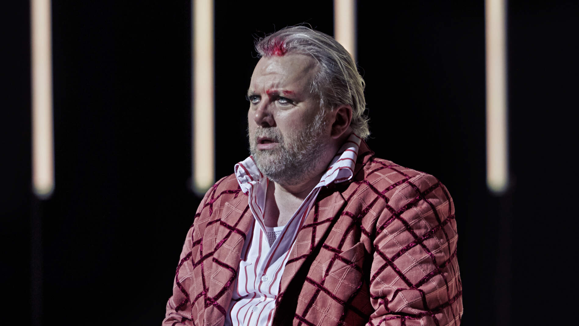Rigoletto sitter och stirrar. Han har blod i håret och över ena ögonbrynet. Han bär en rutig kostym.
