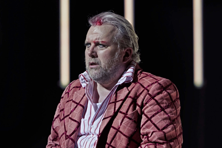 Rigoletto sitter och stirrar. Han har blod i håret och över ena ögonbrynet. Han bär en rutig kostym.