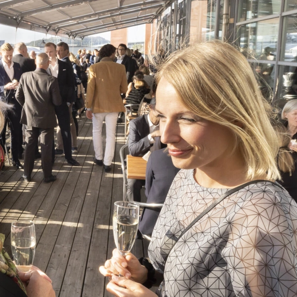 Två personer står och pratar på en restaurangveranda i solen, med varsin champagneglas. Bakom dem står flera andra grupper som minglar och pratar.