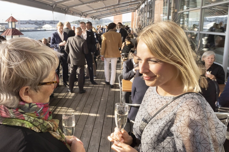 Två personer står och pratar på en restaurangveranda i solen, med varsin champagneglas. Bakom dem står flera andra grupper som minglar och pratar.
