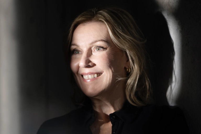 En leende kvinna lutar sig mot en betongvägg med armar i kors. Solljus skiner på hennes ansikte men rummet är annars mörkt.