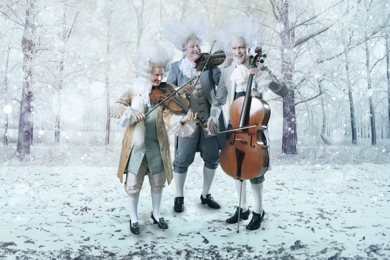 Tre stråkmusiker i barocka kläder och stora vita peruker står och spelar sina instrument på ett snöigt fält omringad av träd.