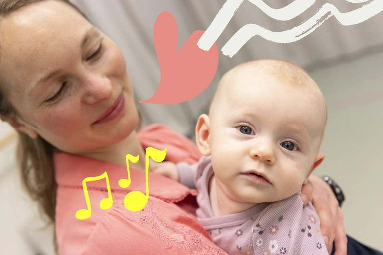 En ung bebis som hålls av sin mamma tittar rakt in i kameran. Hennes mamma tittar på henne och ler.