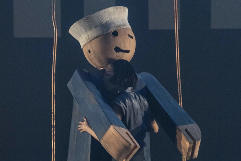 En dansare står och omfamnar en gigantisk träfigur av en blinkande leksakssjöman som är ett huvud längre än henne.