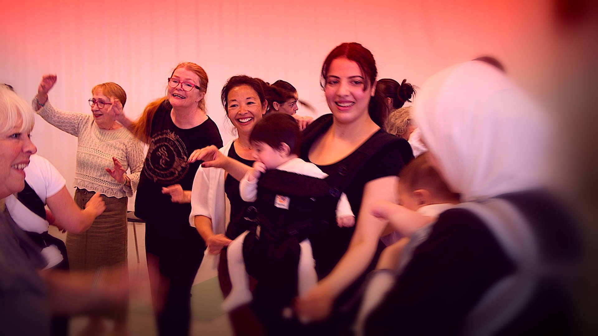 En grupp människor – några äldre, andra mammor med små bebisar i bärselar – dansar tillsammans i en studio.