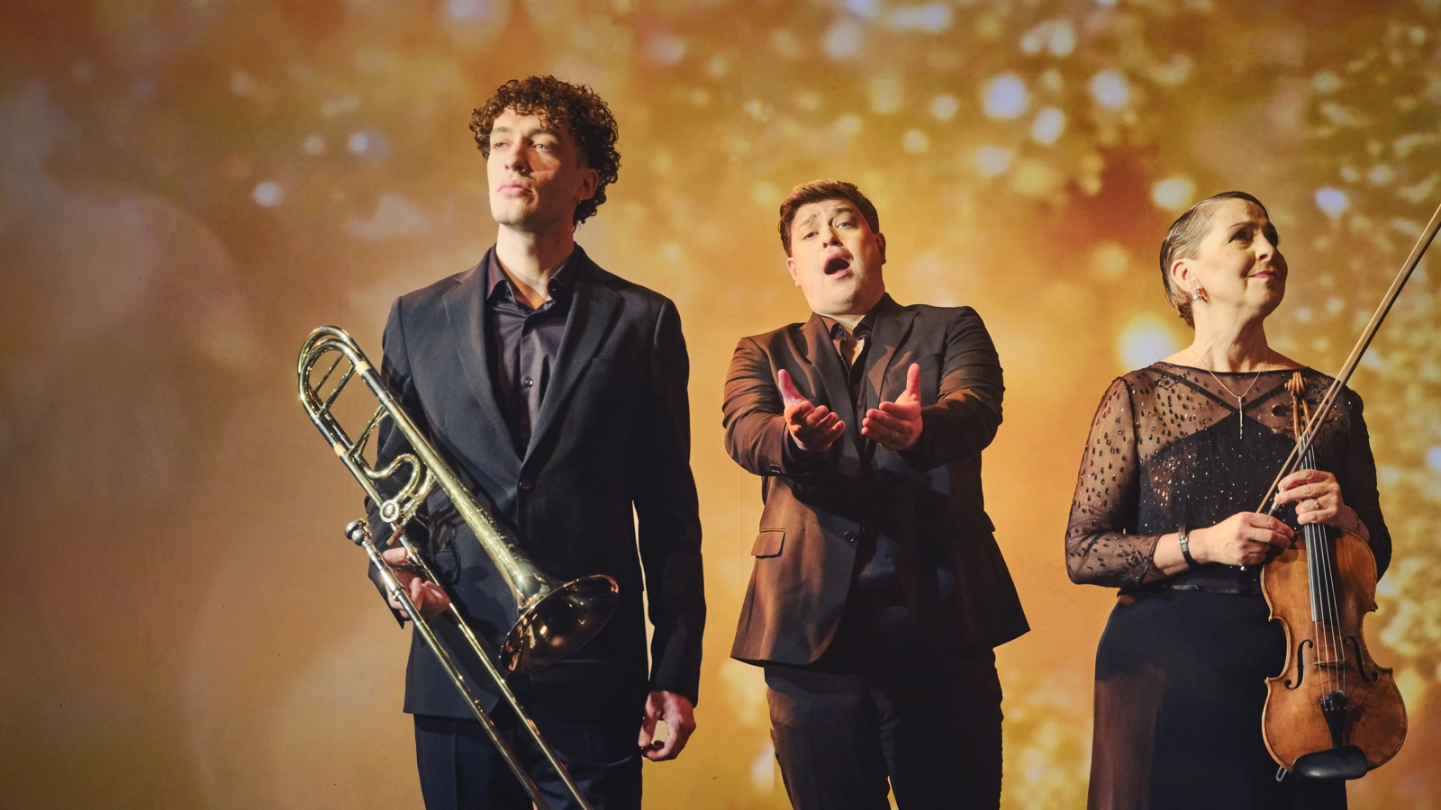 En trombonist, en operasångare och violinist står i svart högtidskläder framför en gul bakgrund. Operasångaren sjunger.