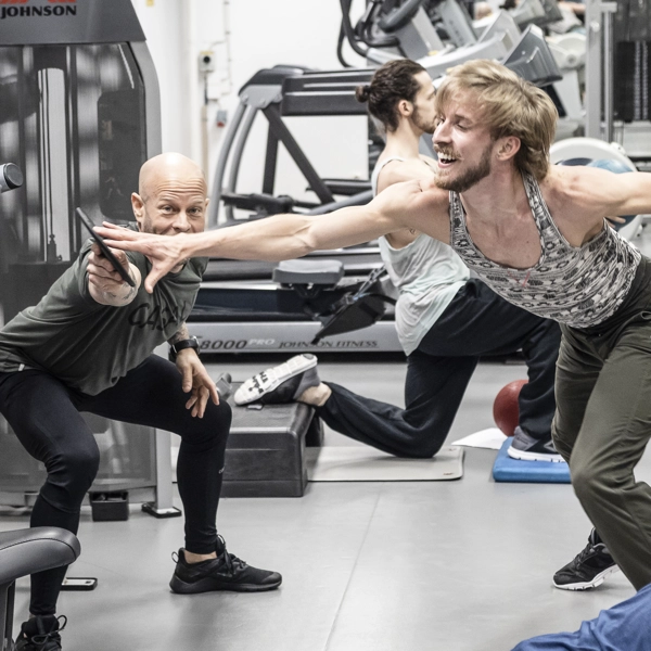 Flera dansare tränar i ett gym. En tränare håller upp en disk som en dansare sträcker handen mot medan ett gummiband är virat runt låret och drar honom i motsatt riktning..