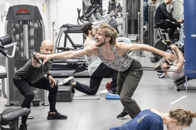 Flera dansare tränar i ett gym. En tränare håller upp en disk som en dansare sträcker handen mot medan ett gummiband är virat runt låret och drar honom i motsatt riktning..
