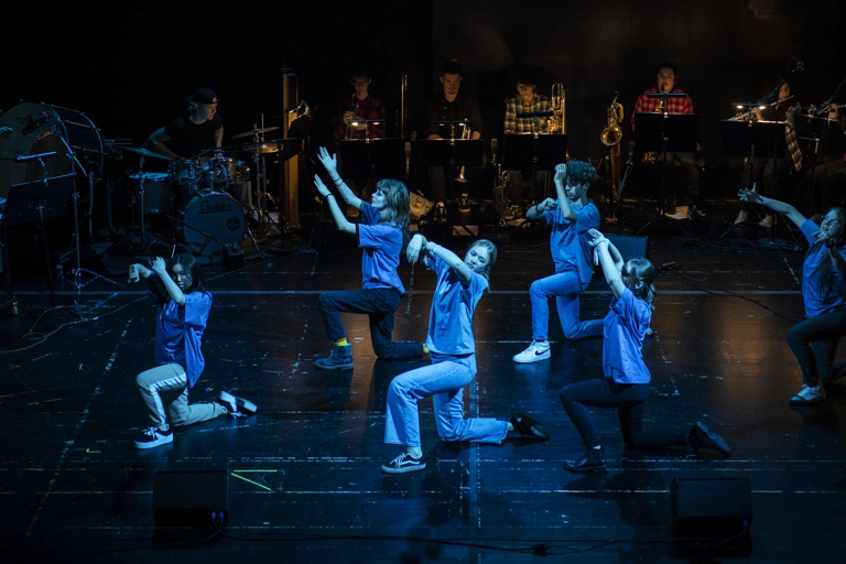 Fem ungdomar står på högra knät och håller armarna ovanför huvudet. De är upplysta i blått. Bakom spelar en rad unga musiker.