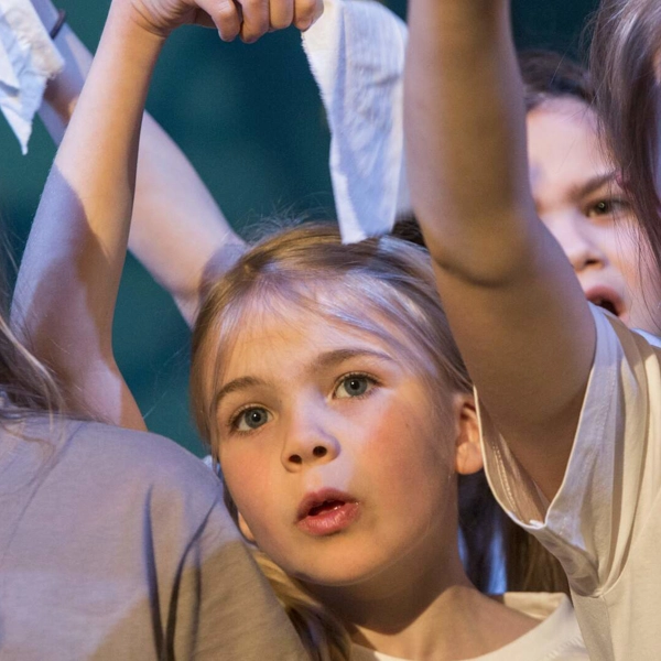 Flera barn i grå eller vita t-shirts står och sjunger och håller upp sin högra arm.
