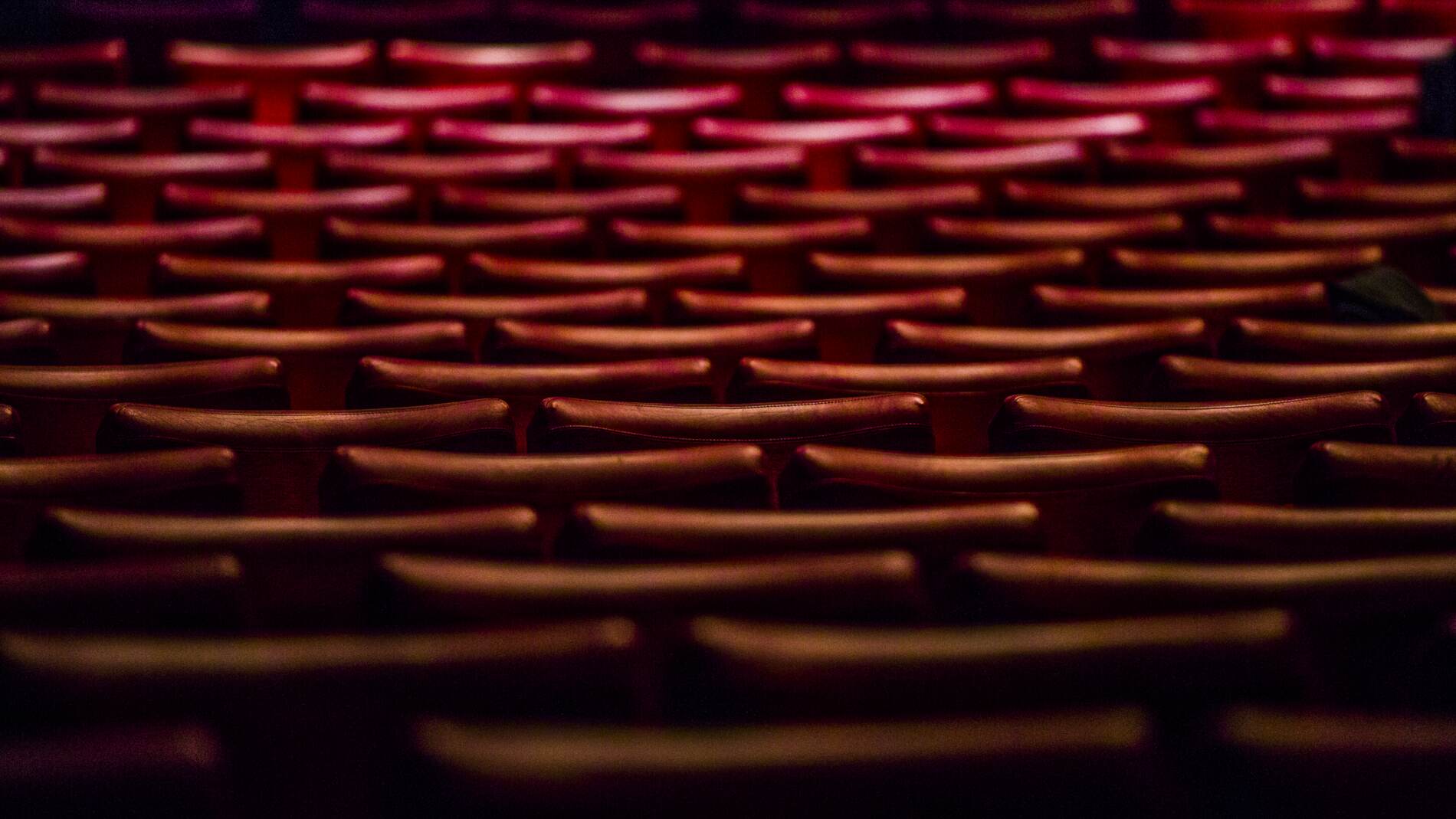Fällstolar av röda sammet står i långa böjda rader i en stor teater auditorium.