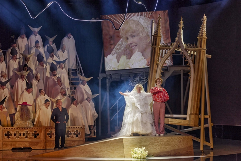 Den unge Hoffmann står på en plattform med Olympia i brudklänning. Oylympia syns på en stor skärm bakom honom, bredvid en läktare fylld med människor i medeltida religiösa skrudar.