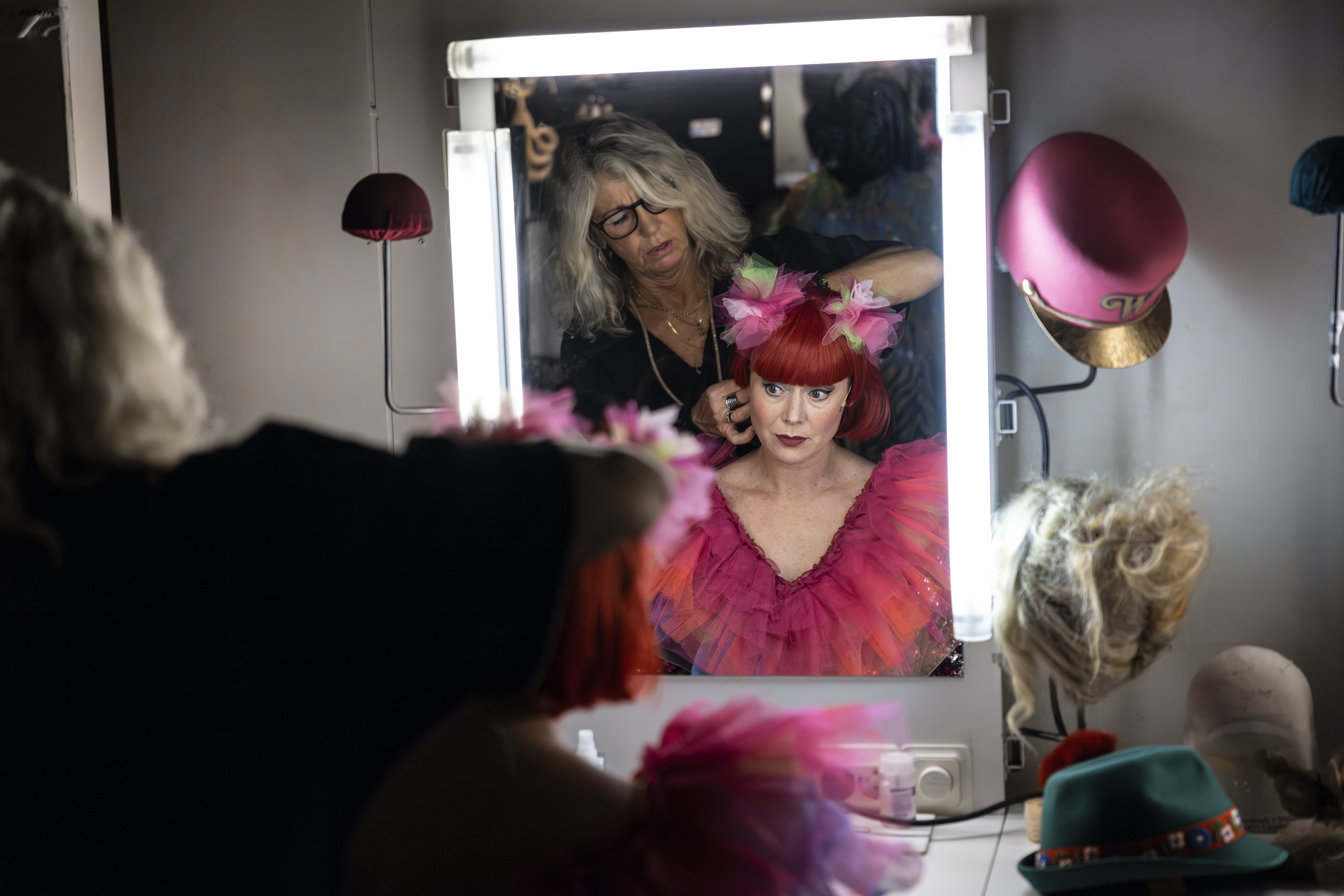 En musikalartist sitter framför en väl upplyst sminkspegel medan makeupartist fixar med hennes röda peruk.