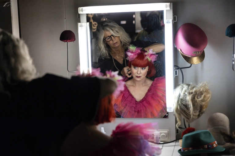 En musikalartist sitter framför en väl upplyst sminkspegel medan makeupartist fixar med hennes röda peruk.