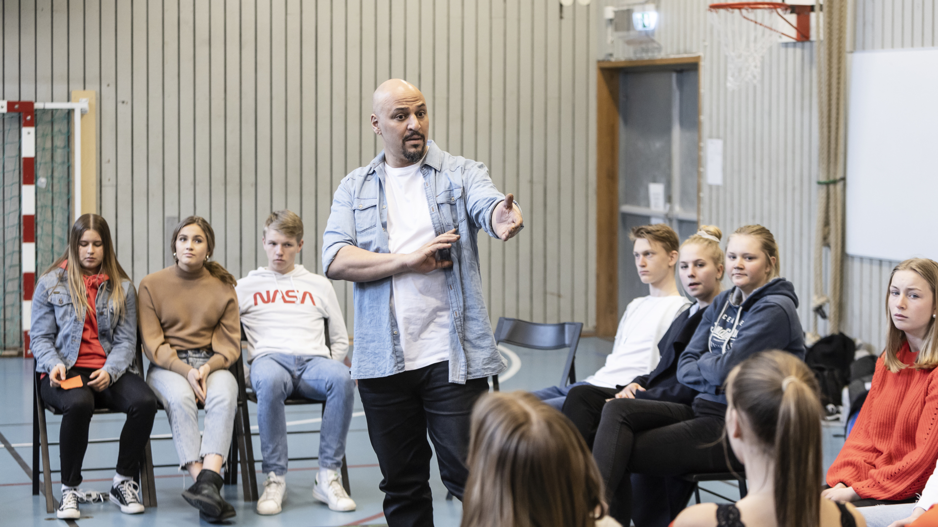 En operasångare står och pratar i ett skolgym, bland en grupp elever som sitter på stolar runt honom.