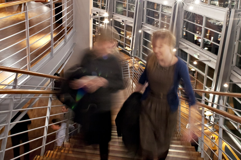 En långexponeringsbild av två personer som går uppför en trätrappa i en modern teaterfoajé.