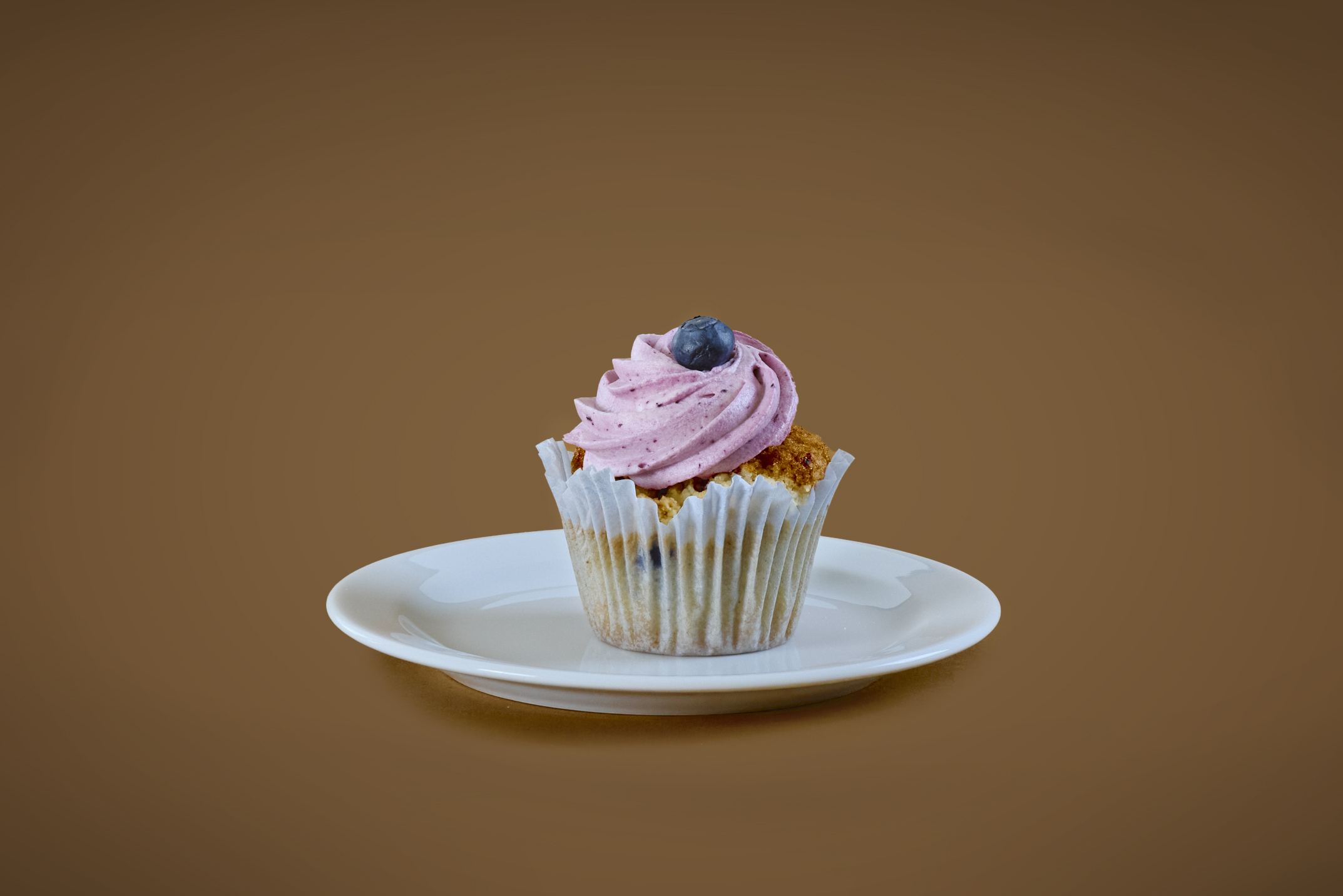En cupcake med rosa frosting och ett blåbär ovanpå ligger på en liten tallrik.