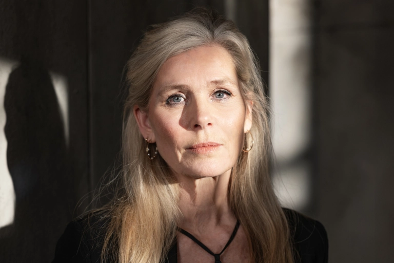 En kvinna lutar sig tillbaka mot en grå vägg. Ljus skiner på ansiktet och delar av väggen, men rummet är annars i skugga.