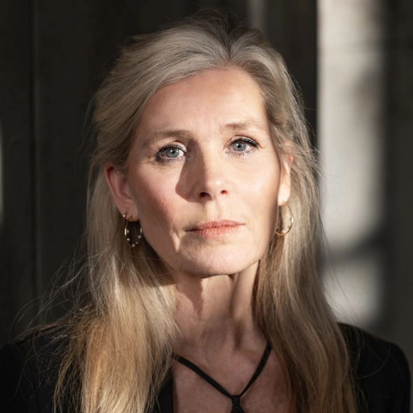 En kvinna lutar sig tillbaka mot en grå vägg. Ljus skiner på ansiktet och delar av väggen, men rummet är annars i skugga.