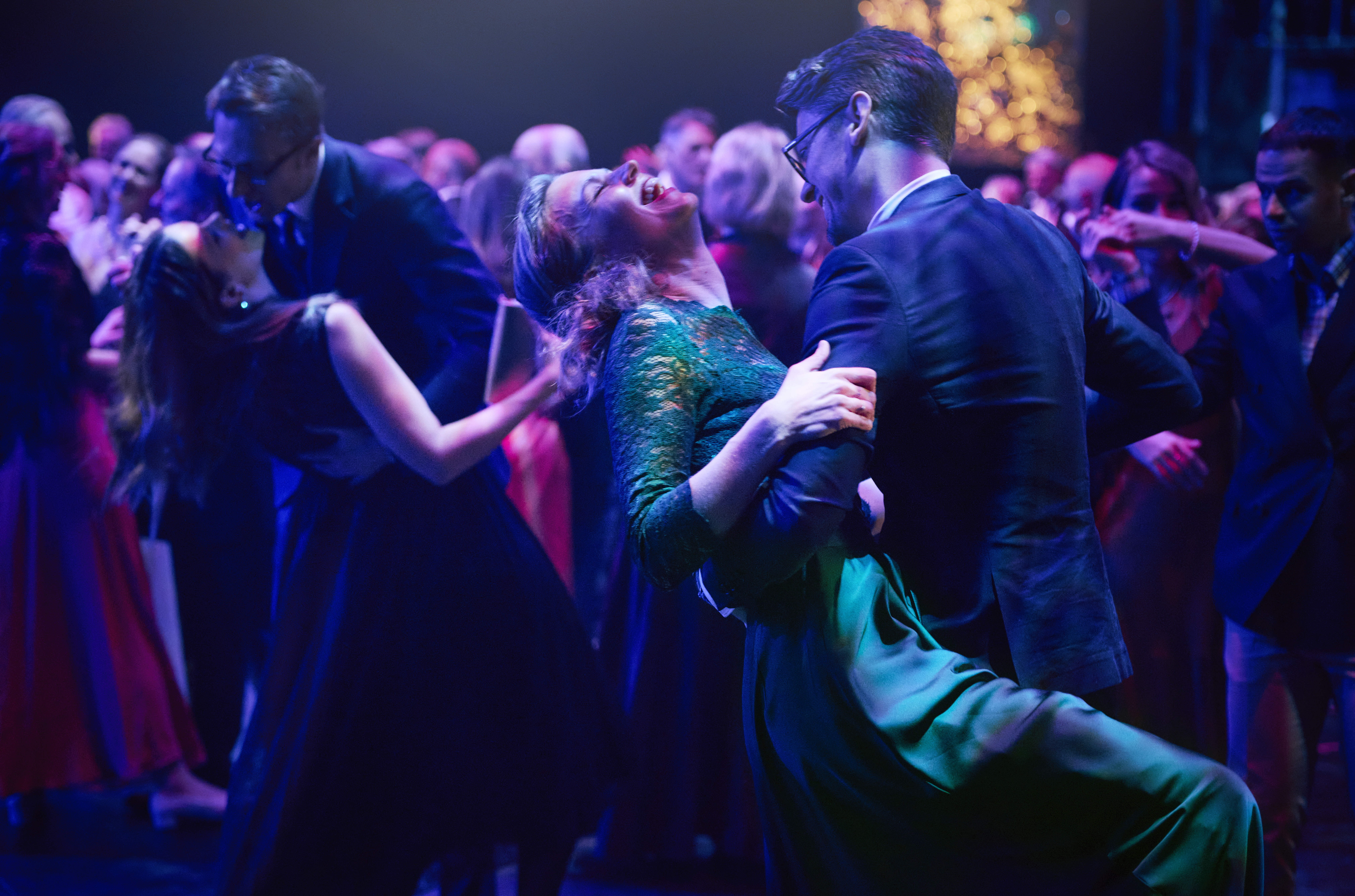 Många par dansar i en balsal under lila ljus. En av paret i förgrunden lutar sig bakåt medan hennes partner håller i henne.