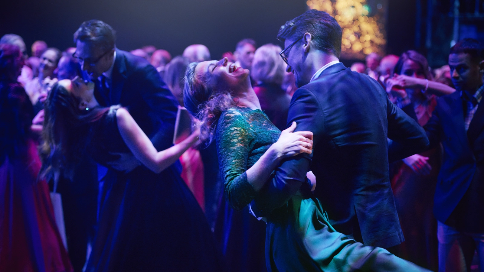 Många par dansar i en balsal under lila ljus. En av paret i förgrunden lutar sig bakåt medan hennes partner håller i henne.