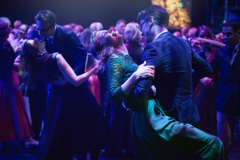 Många par dansar i en balsal under lila ljus. En av paret i förgrunden lutar sig bakåt medan hennes partner håller i henne.