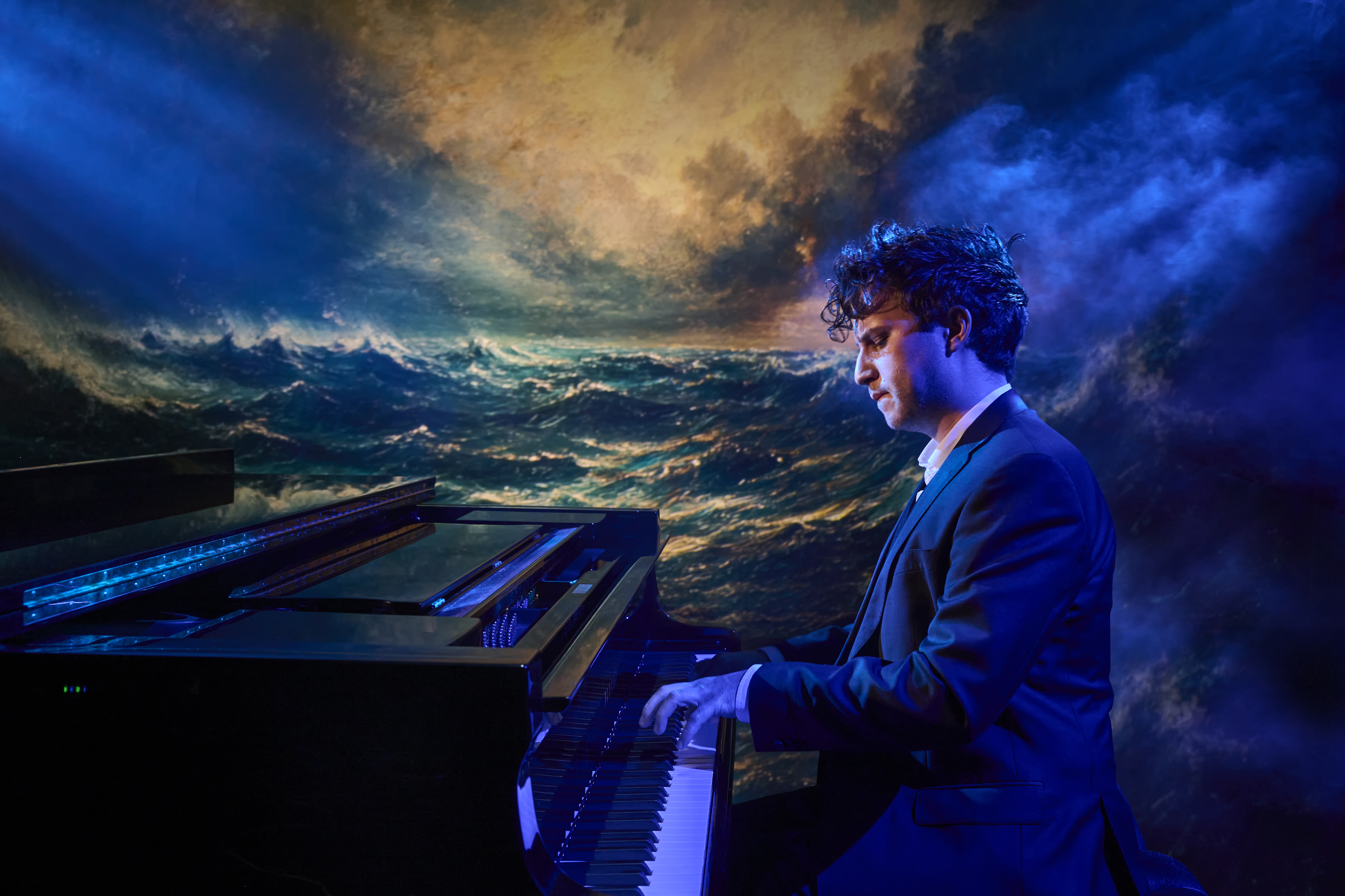 En pianist sitter och spelar piano. Han är lyst i svala färger, och på kulissen bakom finns en bild av ett stormigt hav.