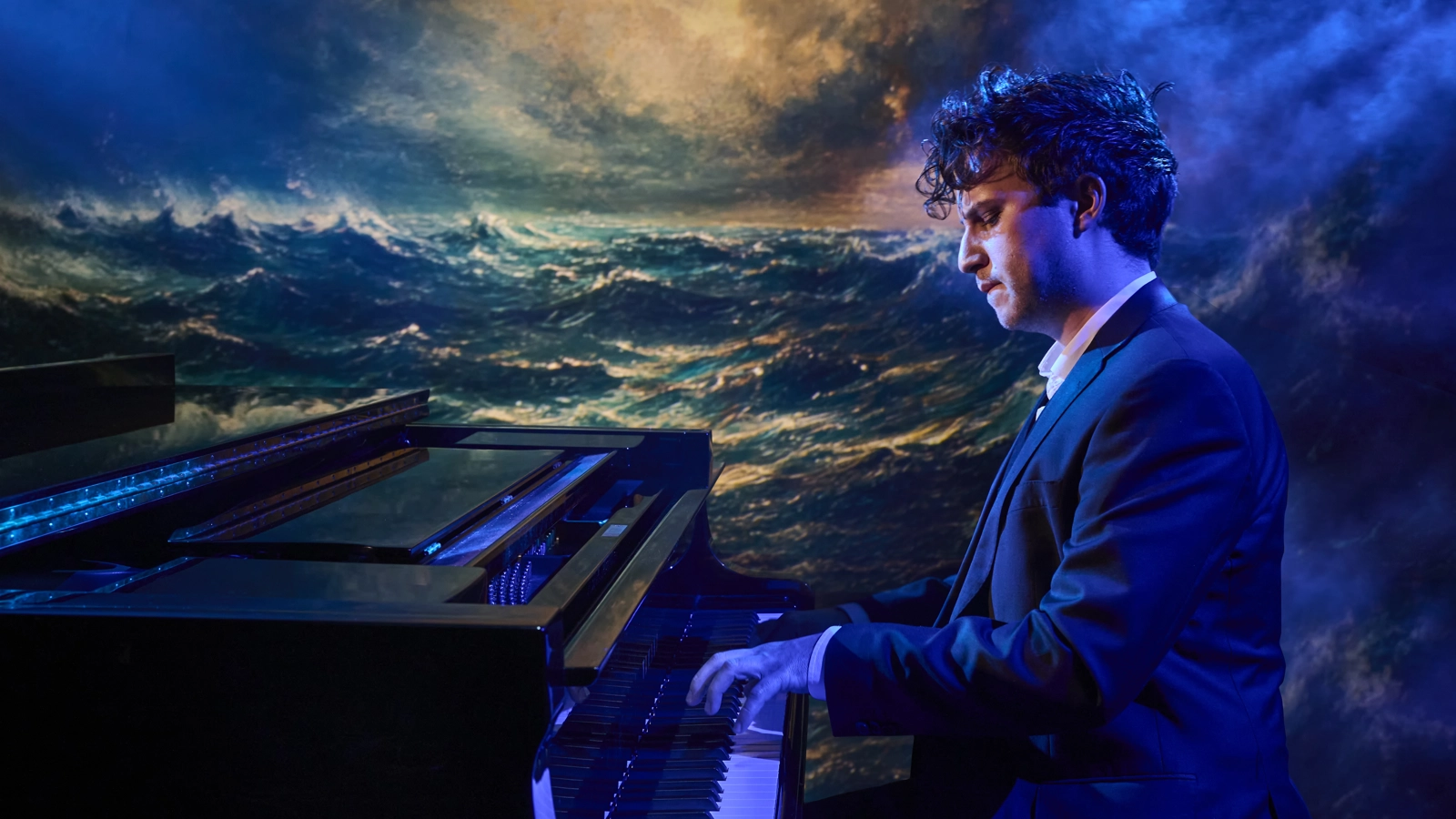 En pianist sitter och spelar piano. Han är lyst i svala färger, och på kulissen bakom finns en bild av ett stormigt hav.