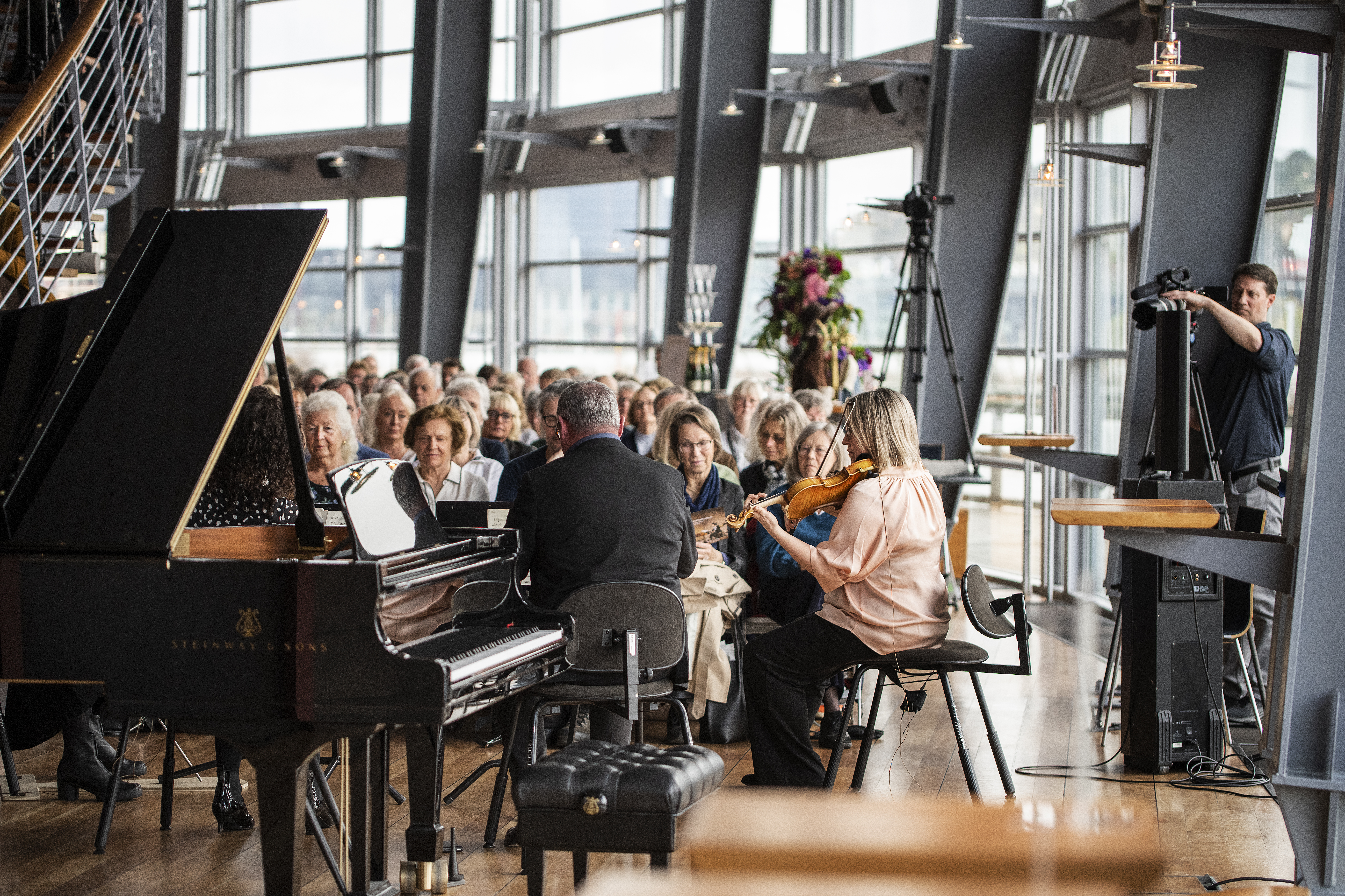 Två musiker sitter och spelar i en väl upplyst teaterfoajé medan en publik tittar på. En flygel står i förgrunden.