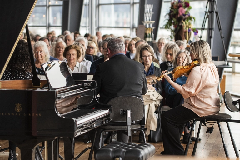 Två musiker sitter och spelar i en väl upplyst teaterfoajé medan en publik tittar på. En flygel står i förgrunden.