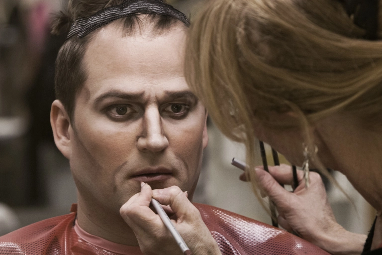 En skådespelare tittar in i en spegel medan en makeupartist sminkar honom i ansiktet.