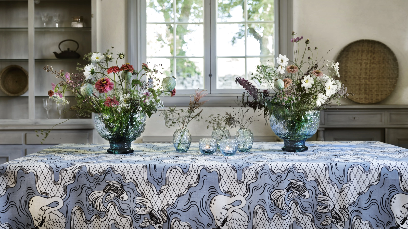 Två stora glasskålar fyllda med blommor på ett bord täckt med mönstrad linneduk. Mellan dem står 5 mindre glasskålar.