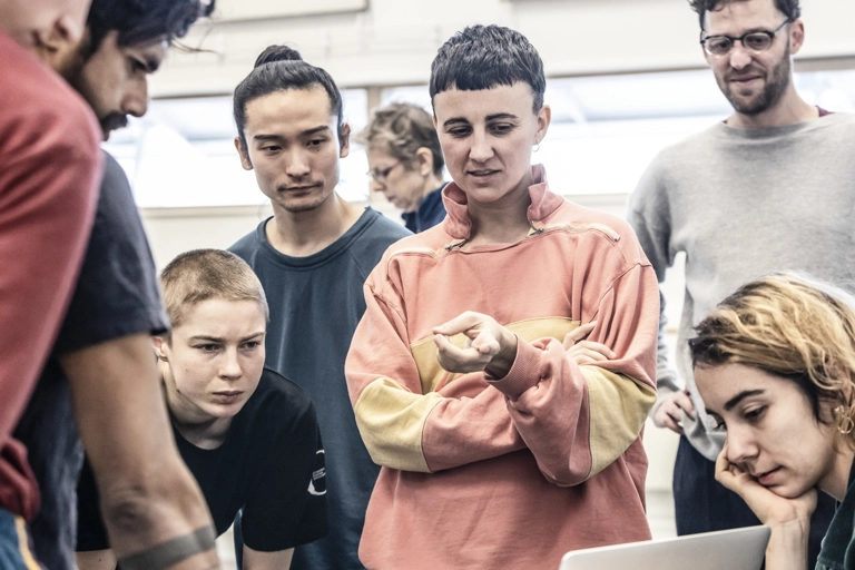 En grupp i en ljus dansrepetitionsstudio står tillsammans och tittar på en öppen laptop.