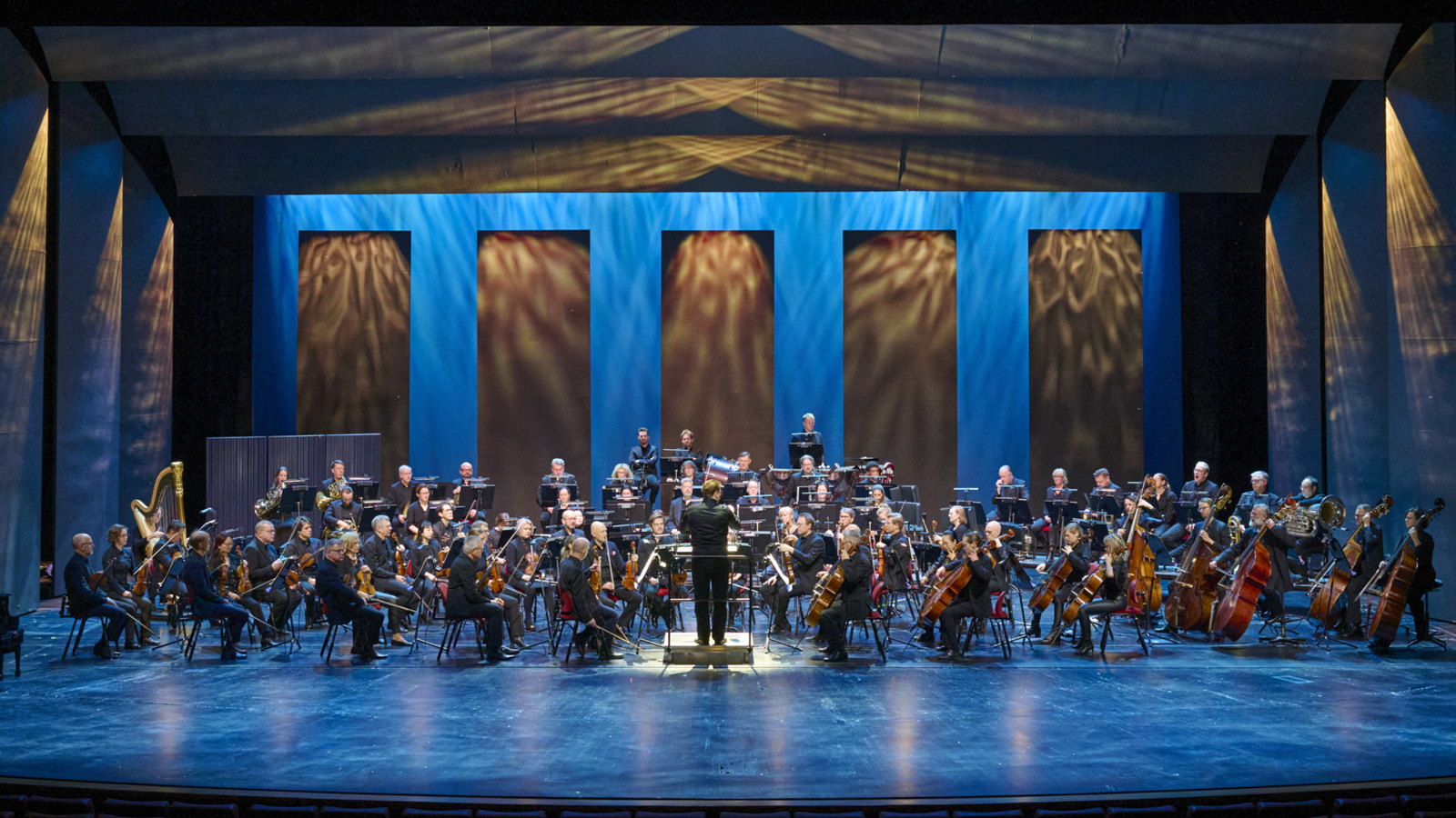 En stor orkester och dirigent är på en upplyst teaterscen.