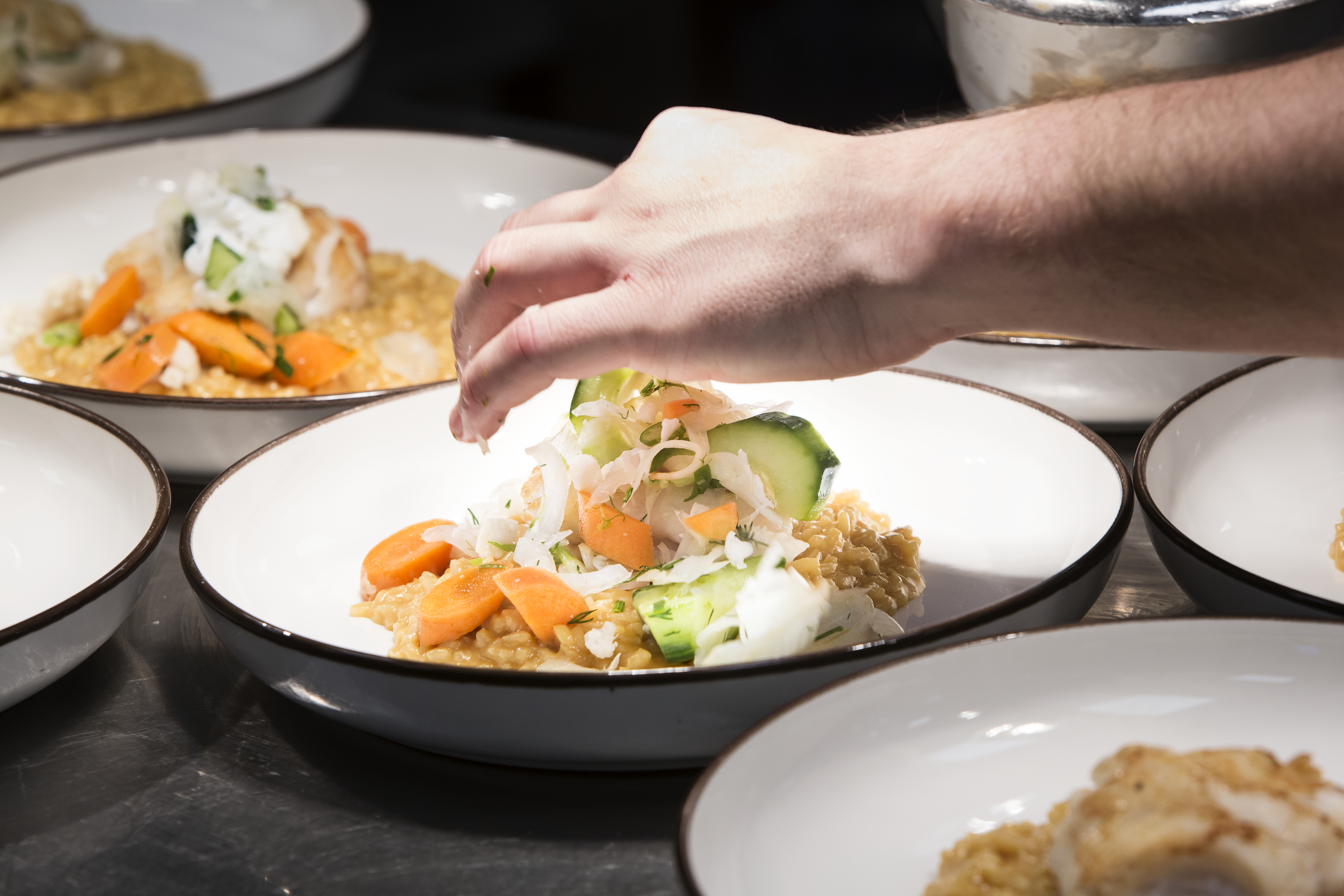 En hand garnerar en rätt som står på tallrik under serveringslampor på en restaurang. Liknande rätter är uppställda i närheten.