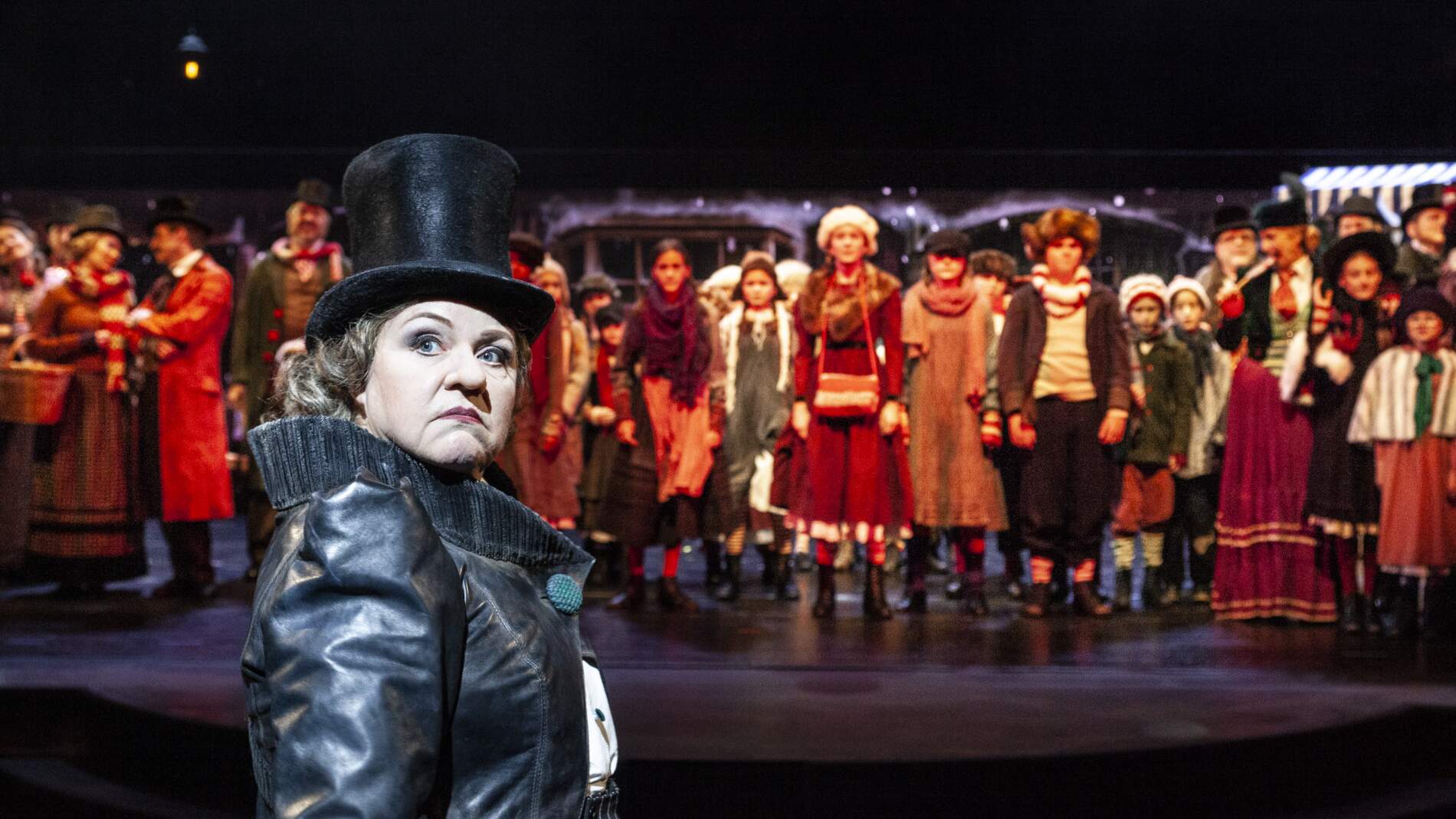 Scrooge står och tittar tillbaka över sin högra axel. Bakom henne står en rad människor i vinterkläder från sekelskiftet.