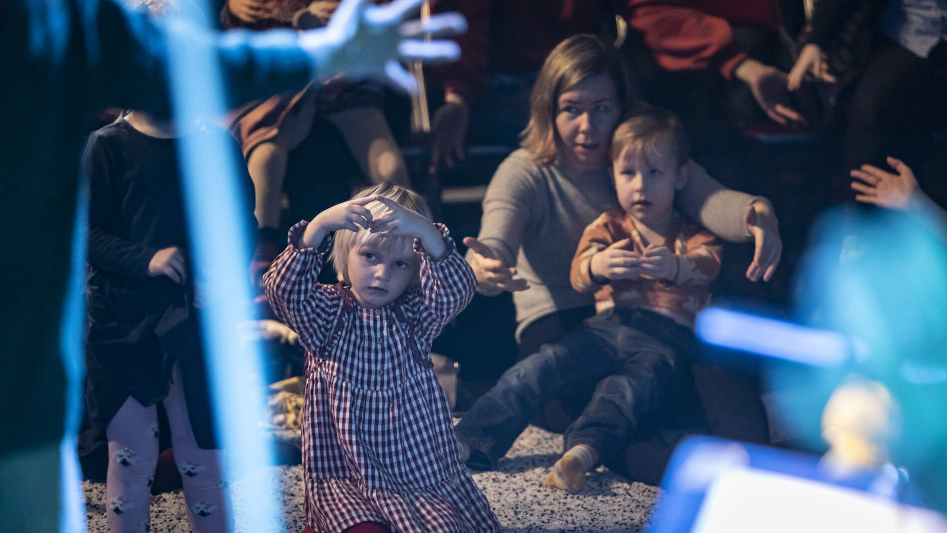 Barn och vårdnadshavare sitter på en tjock matta och tittar på en föreställning. Några personer sitter på stolar bakom.