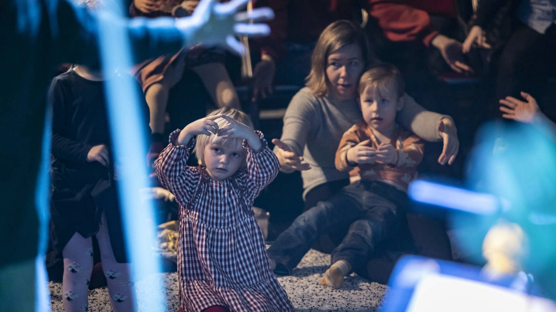 Barn och vårdnadshavare sitter på en tjock matta och tittar på en föreställning. Några personer sitter på stolar bakom.